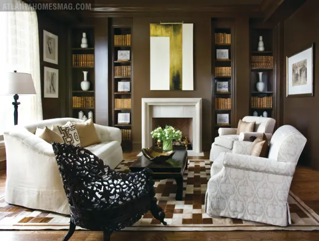 Brown living room with white tufted sofa, ornate dark wood chair, patterned armchairs, fireplace, and built-in bookshelves filled with books.