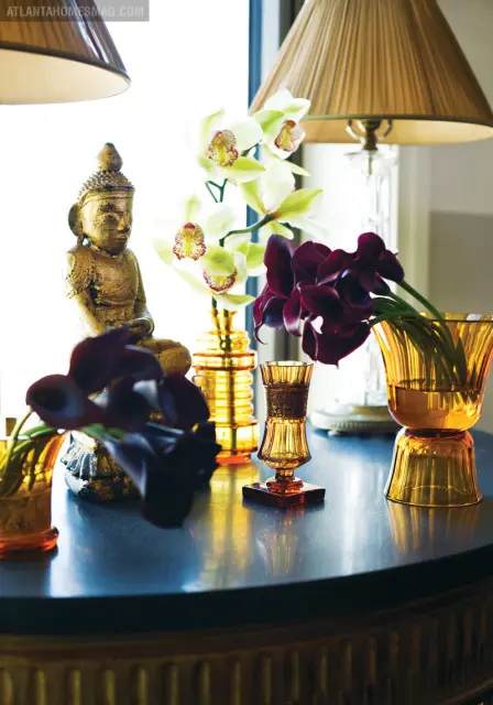 A golden Buddha statue sits on a dark table next to amber glass vases holding white orchids and dark calla lilies.