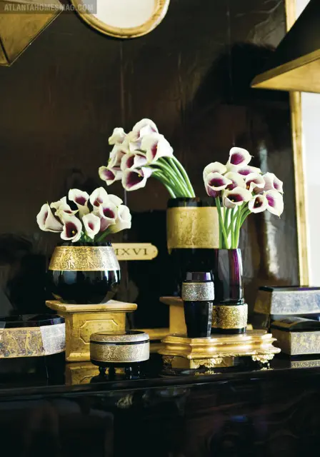 A collection of black vases with gold detailing holds white calla lilies with dark purple centers. The vases sit on ornate gold pedestals against a dark, textured wall.