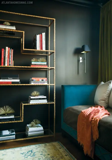 Dark room with a gold geometric bookshelf filled with books and decorative objects, a teal upholstered headboard, and a textured throw blanket.