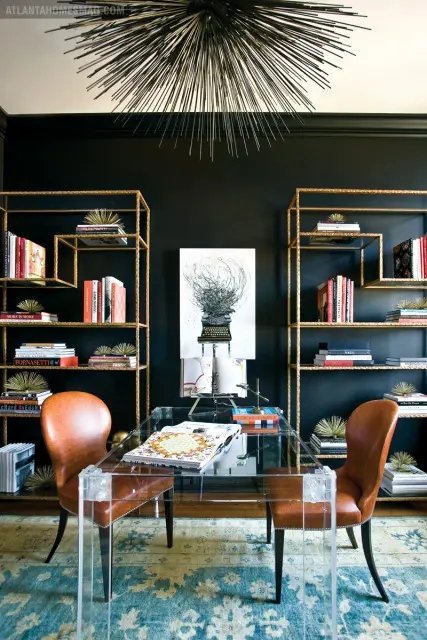 Black office with a clear acrylic desk, two brown leather chairs, gold shelving units filled with books, and a spiky black chandelier.