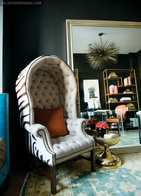 Tufted high-back armchair with zebra print accents and an orange throw pillow, next to a gold hourglass side table with a vase of red roses.