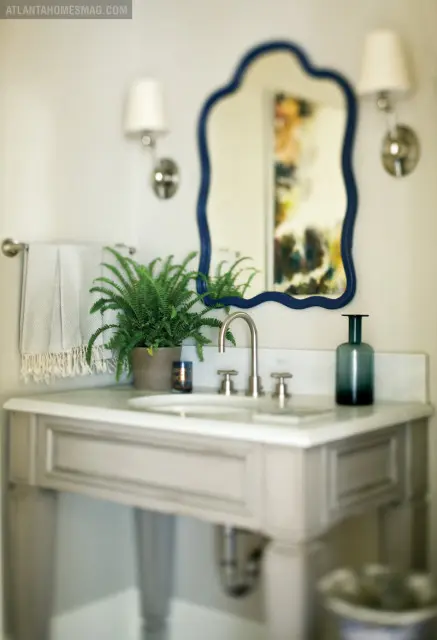 Powder room with a gray vanity, marble countertop, white sink, brushed nickel faucet, and a scalloped navy mirror. A potted fern and a dark green glass vase sit on the counter.