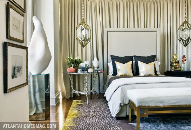 Bedroom with a large upholstered headboard, dark pillows with gold patterned accents, and a white fur bench. Neutral curtains hang behind the bed, and a geometric rug covers the floor.