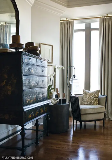 Living room corner with dark lacquered chest, armchair with patterned pillow, and tall windows with beige curtains.