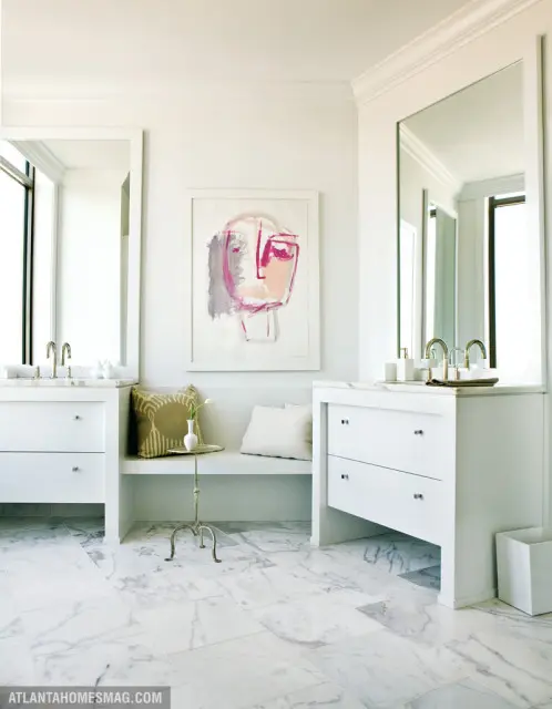 White bathroom with marble tile floors, two white vanities with brass faucets, and a built-in bench with abstract art above