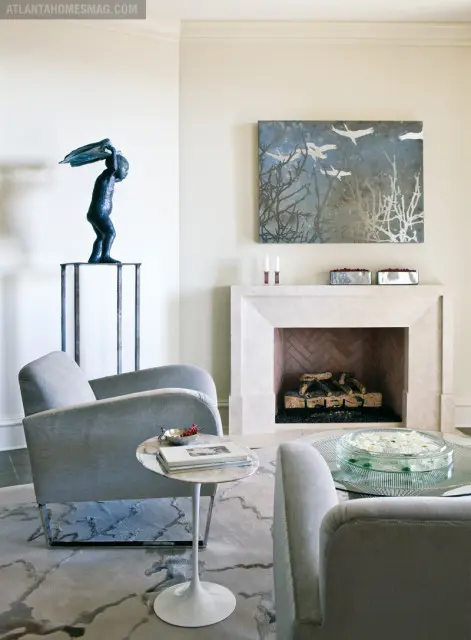 Living room with two gray upholstered armchairs, a marble-topped side table, and a stone fireplace with herringbone brick surround. Above the fireplace hangs abstract art of birds and branches.