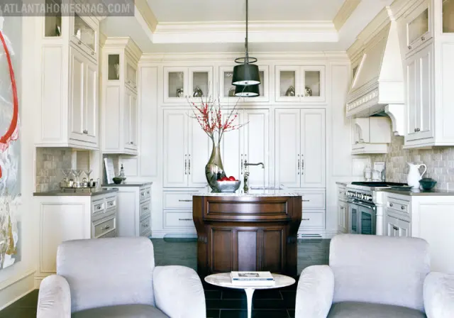 Cream kitchen with white custom cabinetry, marble island with dark wood base, and stainless steel range. Two upholstered chairs face a small marble tulip table.