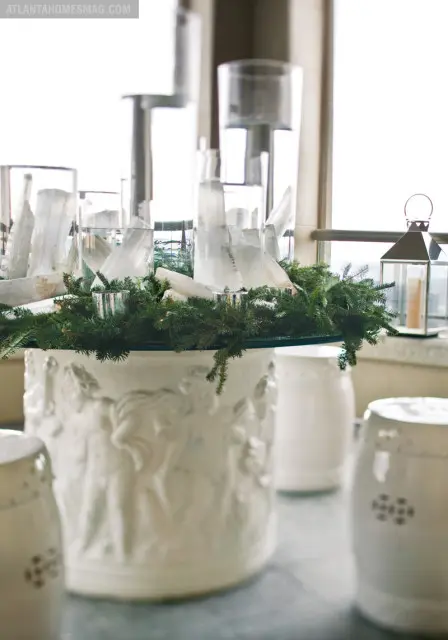 White pedestal with bas-relief figures, topped with a glass surface displaying a festive arrangement of evergreen branches, clear glass cylinders, and white crystal shards.