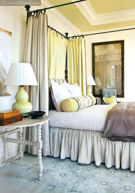 Bedroom with a four-poster bed draped in yellow and striped fabric, a quilted white bedspread, and a patterned bolster pillow. A distressed mirror hangs on the wall above a lamp.