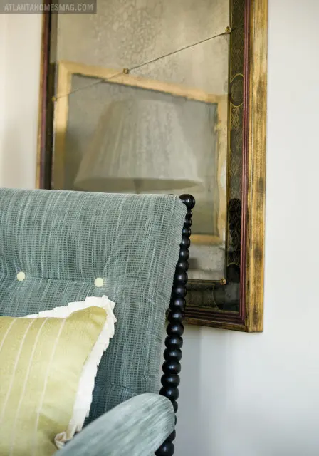 Close-up of a light blue upholstered armchair with tufted buttons and a yellow striped pillow, next to an antique mirror with a distressed finish and ornate gold frame.