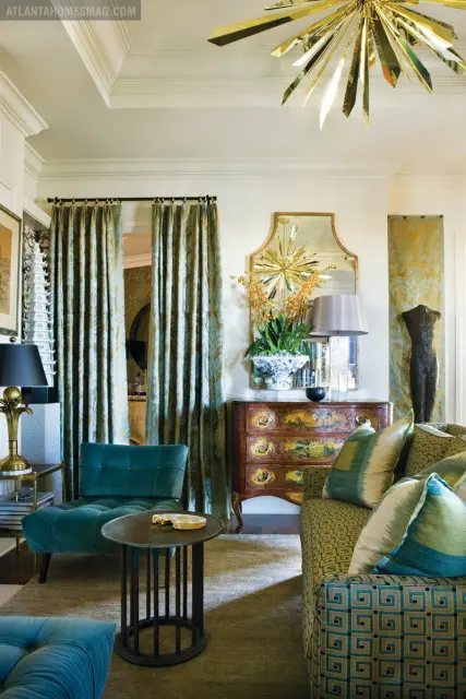 Living room with teal velvet armchair, patterned sofa, ornate chest of drawers, and a gold sunburst mirror above