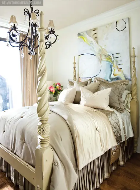 Bedroom with a cream-colored four-poster bed, tufted comforter, satin pillows, and a crystal chandelier. A large abstract painting hangs above the bed.