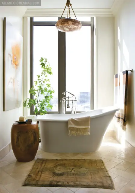 Freestanding white bathtub with chrome fixtures, a beige towel draped over the side, and a plant in a wooden stool beside it. A patterned rug lies on the tiled floor in front of the tub.