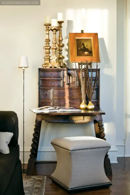 Antique wooden desk with ornate gold candlesticks, a framed painting, and decorative feathers in gold vases. An open book rests on the desk, with a floor lamp and upholstered ottoman nearby.