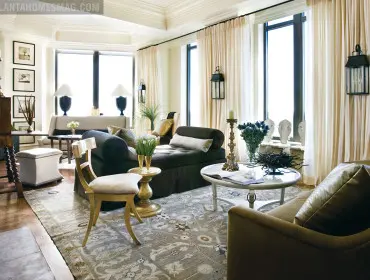 Living room with dark upholstered sofa, chaise lounge, and accent chair, featuring a large patterned rug, ornate candelabra, and floor-to-ceiling cream curtains.