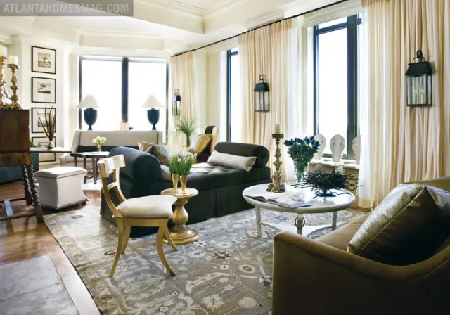 Living room with dark upholstered sofa, chaise lounge, and accent chair, featuring a large patterned rug, ornate candelabra, and floor-to-ceiling cream curtains.