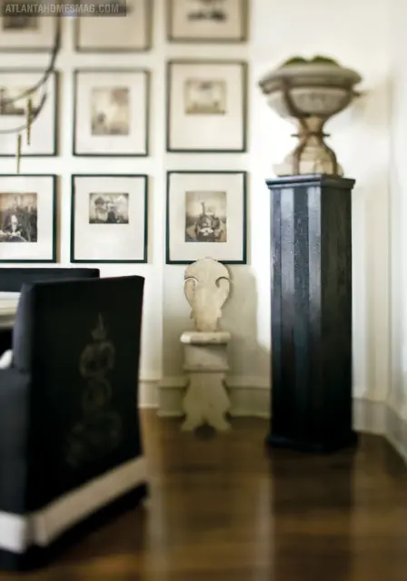 Dining room with dark upholstered chairs, a gallery wall of framed vintage photographs, and a tall black pedestal topped with a stone urn.