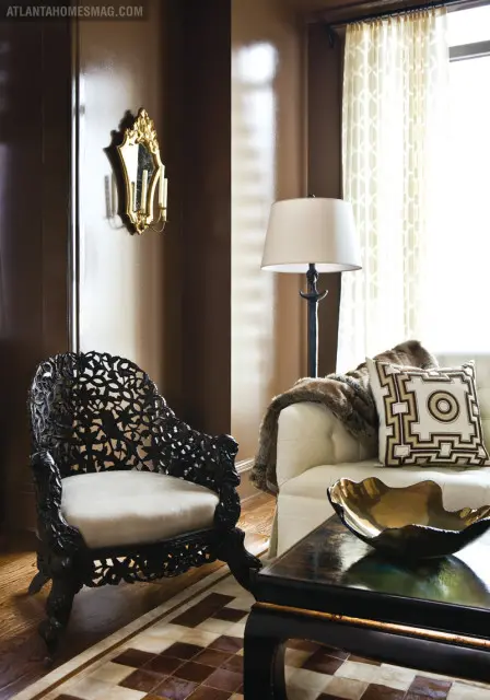 Living room with a dark carved wood armchair, cream sofa with faux fur throw and geometric pillow, and a dark wood coffee table with a gold bowl.