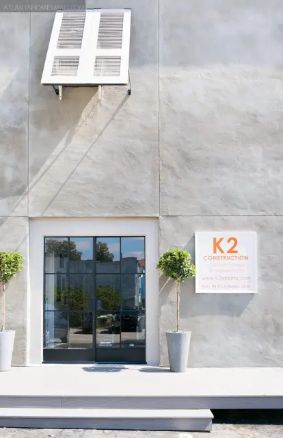 Modern building exterior with gray stucco walls, a white louvered awning, black-framed glass double doors, and two potted trees flanking the entrance.
