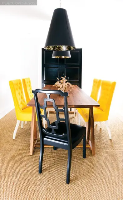 Dining room with a wooden table, black chairs with geometric backs, and yellow tufted chairs. A large black pendant light hangs above the table.