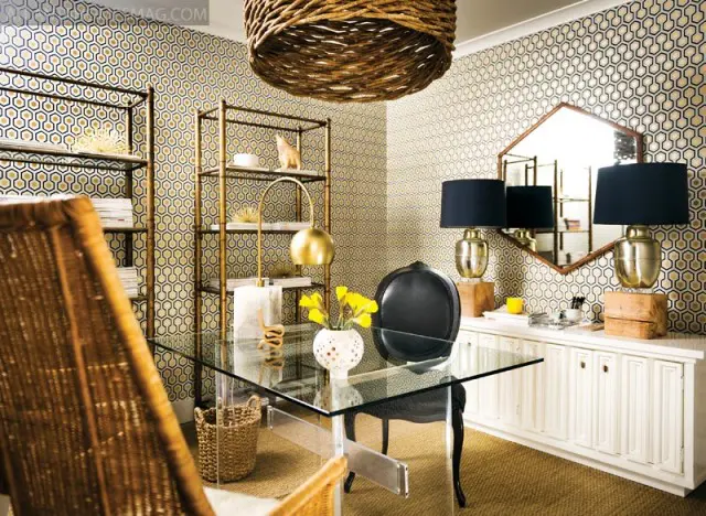 Office with geometric patterned wallpaper, glass desk, rattan chair, brass shelving, and two black lamps on a white credenza.