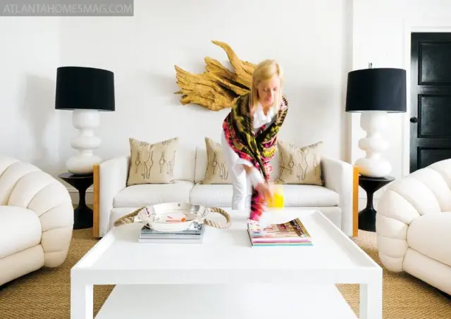 Living room with white sofa, cream upholstered chairs, white coffee table, and black lamps. A woman in a colorful wrap is reaching for a drink.