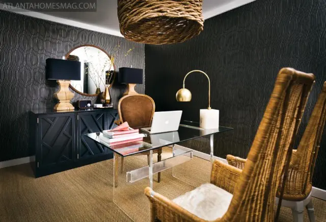 Office with black textured wallpaper, glass desk with laptop, wicker chairs, and a dark wood credenza with a round mirror above it.