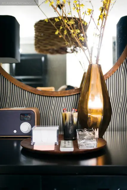 A wooden tray holds a digital radio, a stack of notepads, a remote control, a glass cup with ice, and a horn-patterned cup filled with pens and brushes. A tall, metallic vase with yellow branches sits behind the tray.