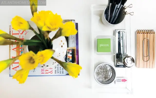 White vase with yellow daffodils on stack of magazines next to acrylic desk organizer with pens, stapler, paper clips, and notepad