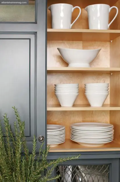 Gray kitchen cabinetry with open shelves displaying white pitchers, a large serving bowl, stacks of small bowls, and stacks of white plates.