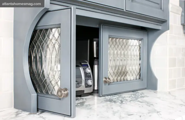 Gray kitchen cabinetry with glass-front doors featuring decorative metal inserts, showing a toaster and coffee maker inside.