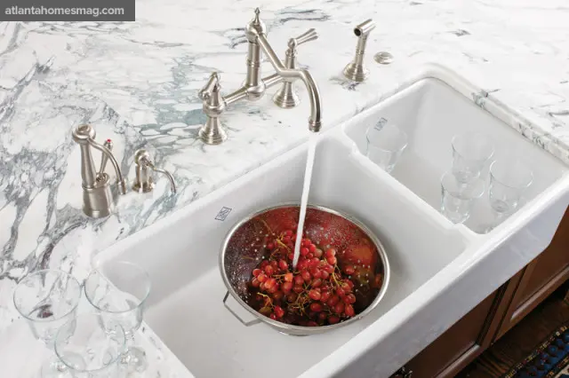 White farmhouse sink with brushed nickel faucet and soap dispenser, marble countertop, and a colander of red grapes under running water.
