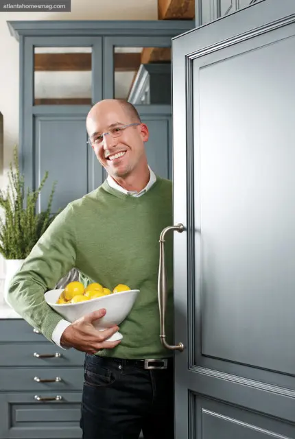 Man in a green sweater holding a bowl of lemons in a gray kitchen with custom cabinetry and wood beams.
