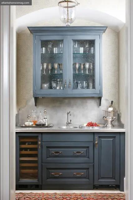 Blue kitchen cabinetry with glass-fronted upper cabinets displaying glassware and silver, a marble countertop with a sink, and a wine fridge.