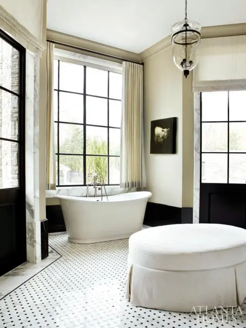 Cream bathroom with a freestanding white tub, black and white mosaic tile floor, white upholstered ottoman, and a glass pendant light.