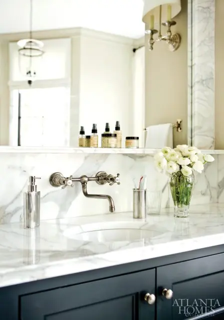 Marble bathroom vanity with dark blue cabinets, a wall-mounted faucet, and a vase of white flowers.