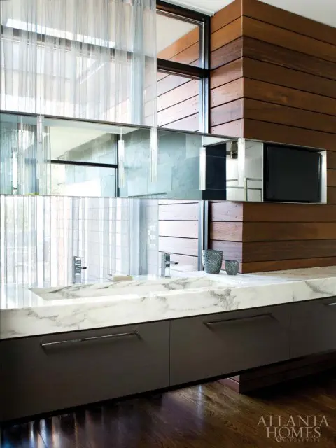 Modern bathroom vanity with a white marble countertop, dark wood cabinetry, chrome fixtures, and a wall-mounted television.