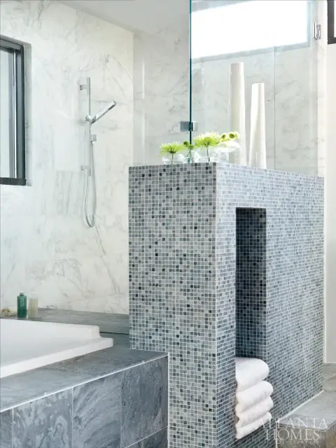 Modern bathroom with white marble walls, a glass shower enclosure, and a mosaic tile pillar with stacked white towels and vases.