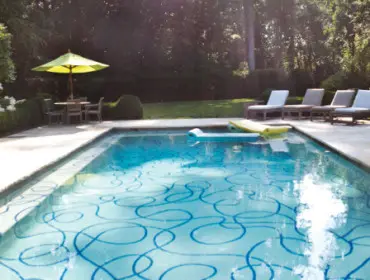 Rectangular swimming pool with blue swirling tile pattern and yellow inflatable float, surrounded by lounge chairs, a table with umbrella, and manicured greenery.
