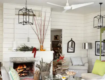 Living room with white shiplap walls, a stone fireplace with a fire burning, and Christmas decorations. Furniture includes two armchairs, a sofa, and a glass-top coffee table.