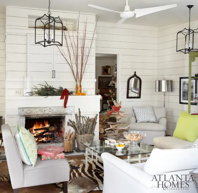 Living room with white shiplap walls, a stone fireplace with a fire burning, and Christmas decorations. Furniture includes two armchairs, a sofa, and a glass-top coffee table.