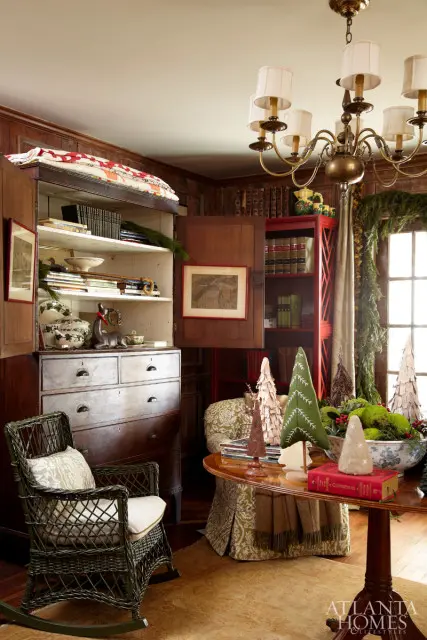Living room with dark wood paneled walls, a dark wicker rocking chair with a patterned cushion, a large antique dresser, and a round wooden table decorated with Christmas trees and moss.