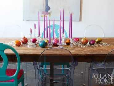 Dining table set for Christmas with purple candles, colorful ornaments, and clear acrylic chairs.