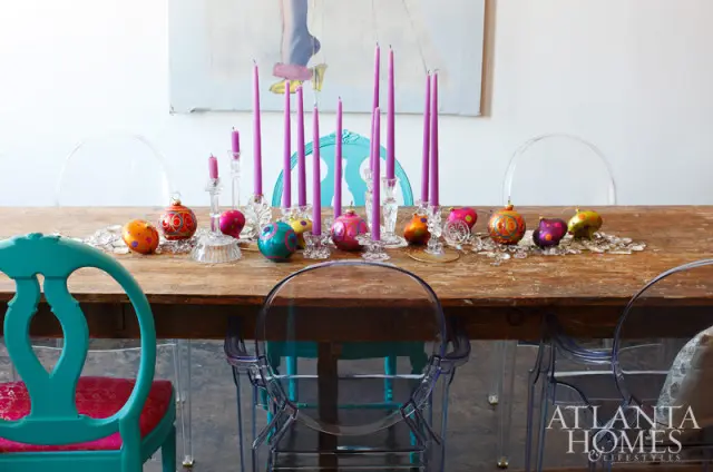 Dining table set for Christmas with purple candles, colorful ornaments, and clear acrylic chairs.