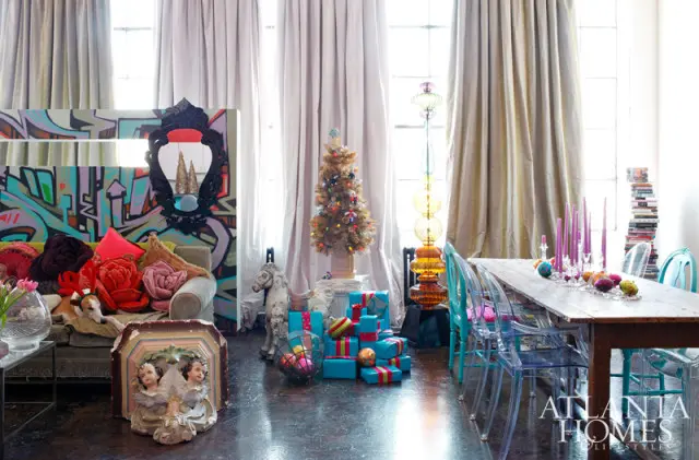 Eclectic living and dining room with graffiti art, a Christmas tree, stacked presents, a dog on a sofa, and a wooden dining table with ghost chairs.