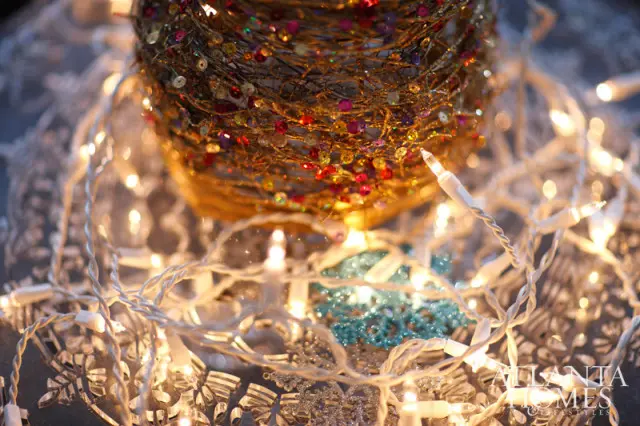 Close-up of a gold, twig-like decorative object adorned with colorful sequins and beads, surrounded by white Christmas lights and silver snowflake ornaments.