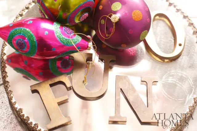 Gold metal letters spelling 'FUN' on a mirrored tray with colorful Christmas ornaments and the Atlanta Homes & Lifestyles logo.