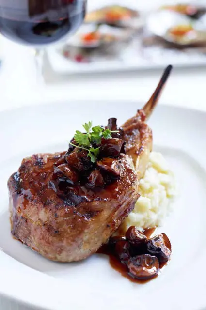 Pork chop with bone, topped with mushroom sauce and parsley, served with mashed potatoes on a white plate.