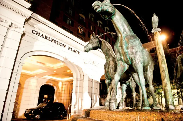 Charleston Place entrance at night with a black car parked under the archway and a bronze horse fountain in the foreground.